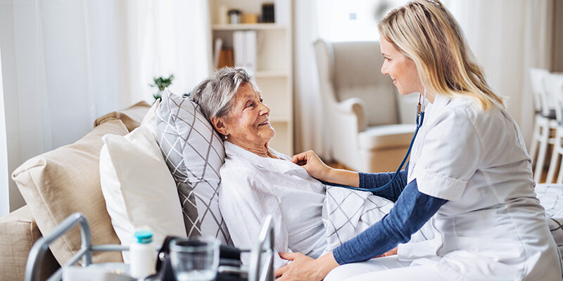 A health visitor examining a sick senior woman lying in bed at home with stethoscope.
